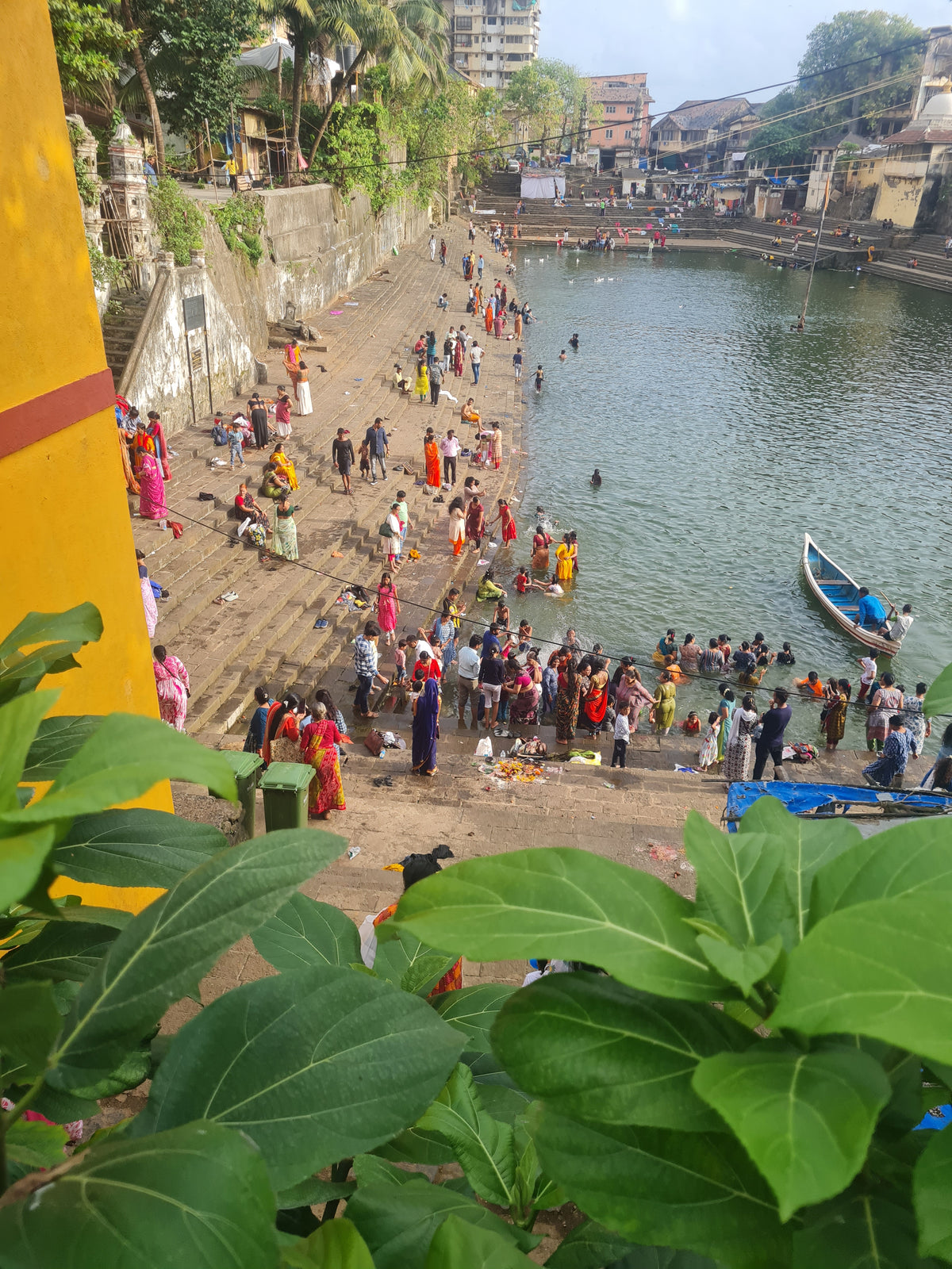 Wider view of Banganga Water Tank in Mumbai, India, showing people gathered along the historic steps by the water, with greenery and surrounding architecture.