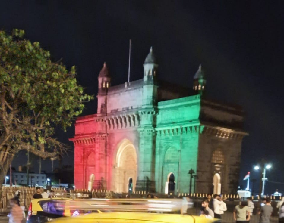 Nighttime photo of the Gateway of India in Mumbai, India, illuminated with red, white, and green vertical light bands resembling the Indian flag, with a yellow taxi blurred in the foreground and people walking nearby.