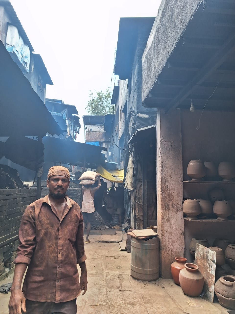 Source photograph of a Dharavi lane in Mumbai, showing a resident walking near pottery shelves and layered urban buildings, used as inspiration for MINA’s painting Resident of Dharavi — Mumbai.